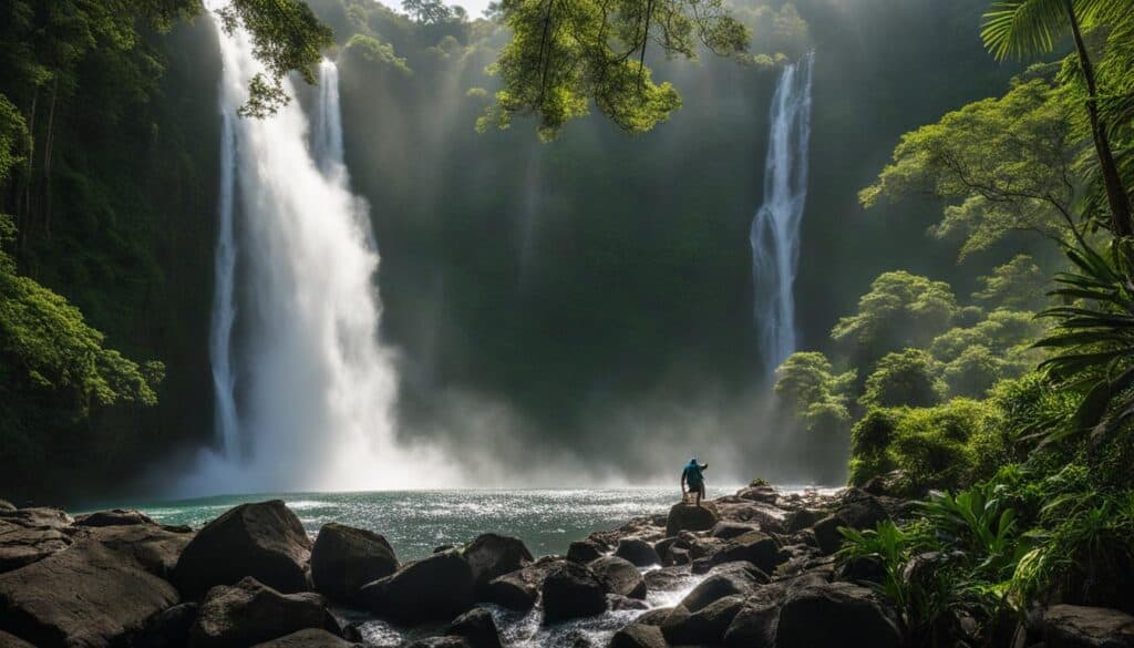 hiker standing in front of Celestine Waterfall, which towers over them and cascades down several tiers of rocks. The lush greenery of the surrounding jungle frames the falls, and mist rises from the water as it crashes into the pool below. 